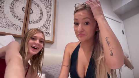 Media: Video of two young Caucasian women with long blonde hair. One woman in a black halter top is adjusting her hair, while the other, in a white top, smiles. Background features a bed with patterned cushions and a white wall.