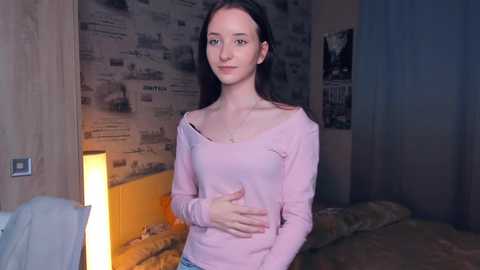 Media: Video of a young, slim, fair-skinned woman with long dark hair, wearing a pink off-shoulder top, standing in a dimly lit bedroom with newspaper wallpaper and a bed.