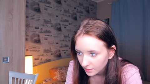 Media: Video of a young woman with long brown hair, wearing a pink top, gazing thoughtfully at a lamp-lit wall covered in vintage newspaper print.