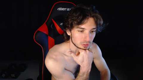 Media: Video of a shirtless, light-skinned man with curly brown hair, sitting in a red and black gaming chair, deep in thought, with a tattoo on his neck.