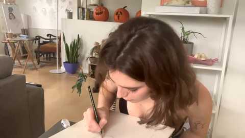 Media: Video of a tattooed woman with shoulder-length dark hair writing in a white notebook at a desk in a cozy, warmly lit room with a potted plant and orange pumpkins on shelves.