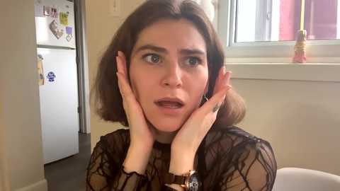 Media: Video of a woman with shoulder-length brown hair, wearing a black lace top, hands on her cheeks, looking surprised, in a kitchen with a fridge and window.