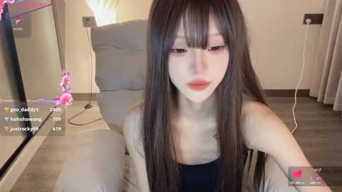 Media: Video of a young East Asian woman with long, straight black hair and pale skin, wearing a black top, sitting on a gray couch in a modern, well-lit room with beige walls and a white lamp.