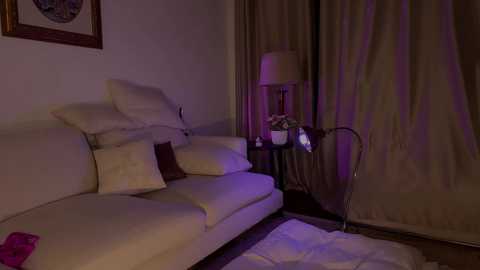 Media: Video of a dimly-lit living room with a cream-colored sofa, a beige lamp, and a decorative plant. Purple ambient lighting adds a cozy atmosphere.