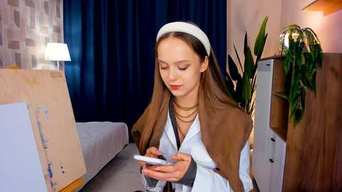 Media: Video of a young woman with long brown hair, wearing a white headband, light-colored coat, and gold necklace, intently looking at her smartphone in a modern, minimalist bedroom with a white bed, dark curtains, and a potted plant.