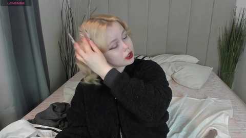 Media: Video of a pale-skinned woman with platinum blonde hair, wearing a black sweater, lying on a bed with white linens, adjusting her hair.