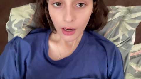 Media: A video of a young Caucasian woman with light skin and brown hair, lying on a bed with floral-patterned pillows, wearing a blue long-sleeve shirt, mouth slightly open, looking surprised or in pain.