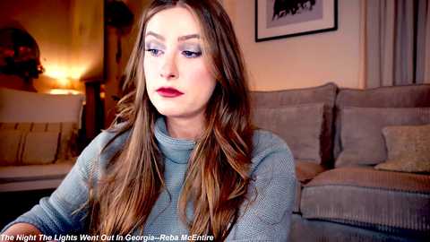 Media: Video of a young woman with long, straight brown hair, wearing a light gray turtleneck sweater, looking contemplative in a cozy living room with beige furniture and warm lighting.