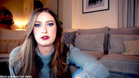 Media: Video of a young woman with long, straight brown hair, blue eyes, and red lipstick, wearing a light blue sweater, sitting on a beige sofa in a warmly lit living room.