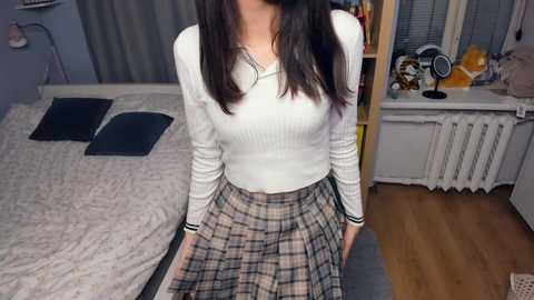 Media: Video of an Asian woman with long dark hair, wearing a white ribbed long-sleeve top and plaid skirt, sitting on a bed in a dimly lit bedroom with a wooden floor.