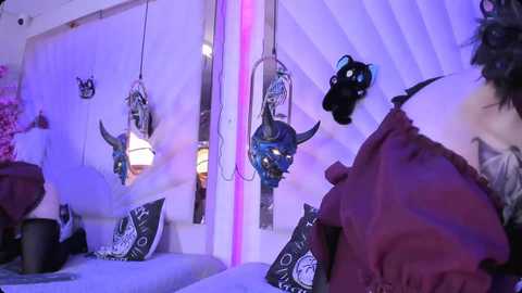 Media: Video of a bedroom with white walls, purple lighting, and a bed adorned with dark purple bedding. Hanging above are black metal bull horns and a black skull.