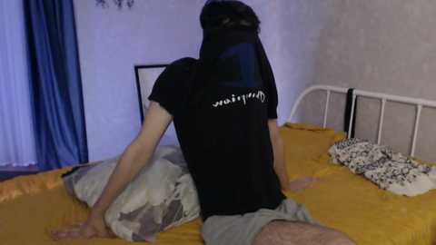 Media: Video of a person with a black mask, wearing a black t-shirt and grey shorts, sitting on a bed with yellow and grey pillows, in a dimly lit room.