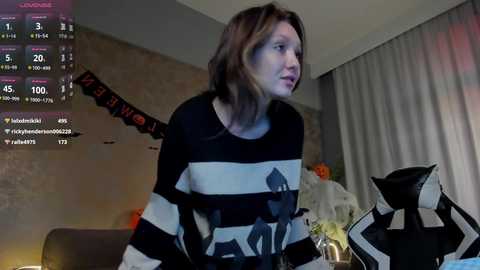 Media: A video of a woman with shoulder-length brown hair in a black-and-white striped sweater, standing indoors near a Halloween-themed backdrop and a black cat statue.