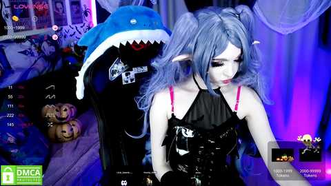 Media: A video of a pale-skinned woman with blue hair in pigtails wearing a black latex dress, standing in a dimly lit room with a large, blue shark costume behind her.