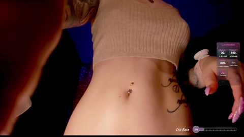 Media: Video of a woman with fair skin and a tattoo on her left hip, wearing a cropped beige top, captured in a dimly-lit room.