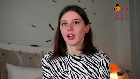 Media: Video of a young woman with fair skin, straight brown hair, and a zebra-patterned shirt, in a bedroom with white bedding and black bird silhouette wallpaper.