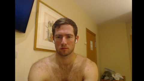 Media: Video of a shirtless, pale-skinned man with short brown hair, wearing white earbuds, standing in a beige room with framed art on the wall and a wooden door.