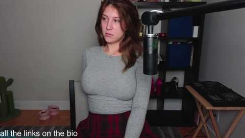 Media: Video of a Caucasian woman with long brown hair in a grey long-sleeve top and red plaid skirt, seated at a desk with a microphone, shelves, and a keyboard visible in the background.