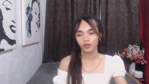 Media: Video of an Asian woman with long brown hair, wearing a white off-shoulder top, standing in a dimly lit room with dark curtains, modern abstract art, and a bouquet of pink and white flowers on the right.