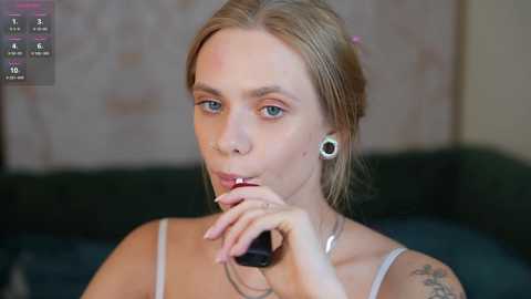 Media: A video of a light-skinned woman with blonde hair, wearing a silver necklace and a gray top, holding a black phone, with a tattoo on her arm, in a blurred indoor setting.