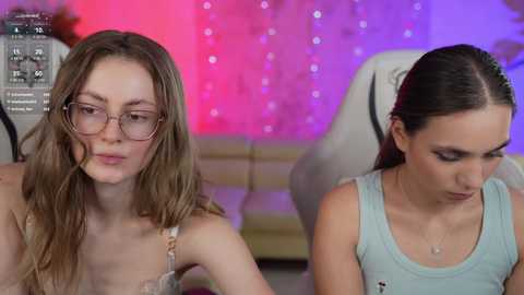 Media: Video of two young women, one with glasses and light brown hair, wearing a white top, and the other with dark hair, wearing a light blue tank top, both looking down. Background features a purple and pink gradient wall.