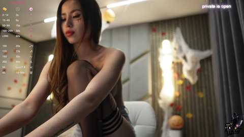 Media: A video of a young Asian woman with long, dark hair, wearing black lingerie, sitting on a bed in a dimly lit room with a TV screen displaying a live stream.