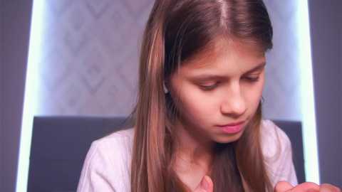 Media: A video of a young girl with long, straight brown hair, wearing a white shirt, looking down, in a modern, grey room with a geometric-patterned wallpaper.