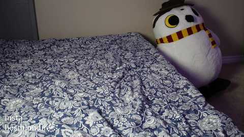 Media: A video of a cozy bedroom with a large, white, plush owl toy partially covered by a blue and white floral bedspread. The owl has yellow eyes and a striped scarf.