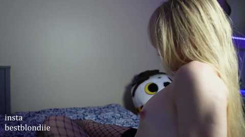 Media: Video of a topless blonde woman with long hair, viewed from behind, standing next to a panda plushie on a bed. The room has a dim, blue light and a white wall. Text reads \"insta bestblondie\" in the bottom left.