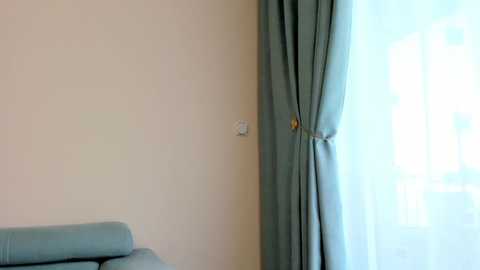 Media: Video of a minimalist room corner featuring a light blue sofa with a rolled-up cushion on the left, a beige wall, and a sheer white curtain partially covering a large window on the right.