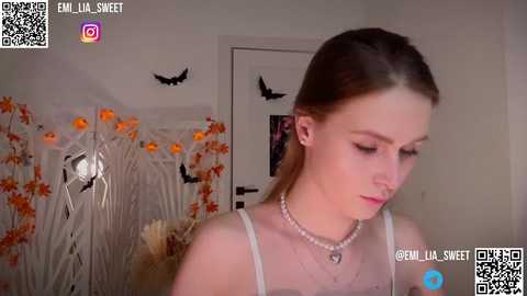 Media: Video of a fair-skinned woman with long brown hair, wearing a white spaghetti-strap top and multiple necklaces, looking down in a cozy room with orange and black decorations.