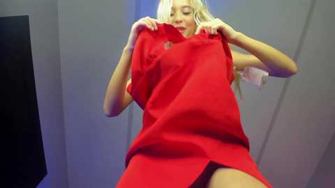 Media: A video of a blonde woman with fair skin, wearing a bright red dress, playfully holding the fabric at her hips. The background is a gray, modern room with a black screen.