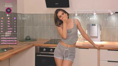 Media: A video of a slender, light-skinned woman with shoulder-length brown hair, wearing a grey crop top and shorts, posing in a modern kitchen, with digital overlay showing streaming details.