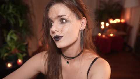 Media: Video of a woman with long brown hair, wearing dark lipstick and a black choker, in a dimly lit room with candles, green plants, and a wooden table.