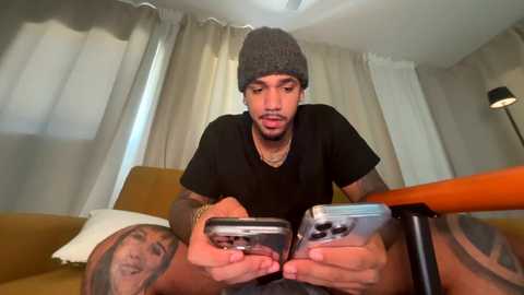 Media: Video of a young man with medium skin tone, wearing a gray beanie and black t-shirt, playing a smartphone game on a bed, surrounded by beige curtains and a mustard-yellow couch.