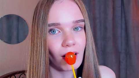 Media: Video of a young woman with long, straight blonde hair and blue eyes, wearing a red lollipop. She appears surprised. Background includes a round mirror and grey curtains.