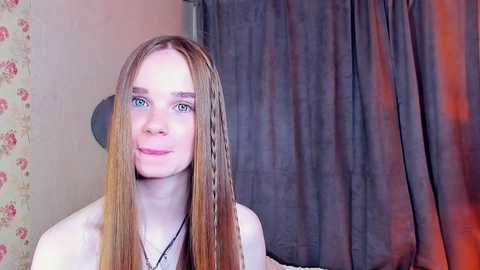 Media: Video of a young Caucasian woman with long, straight, light brown hair parted in the middle, wearing a black necklace. She has blue eyes and a gentle smile. Background shows floral wallpaper, dark curtain, and a lamp with a round shade.