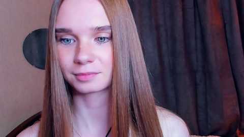 Media: Video of a young woman with long, straight, light brown hair, fair skin, and blue eyes, smiling softly. She wears a black top. Background features dark curtains and a round mirror.