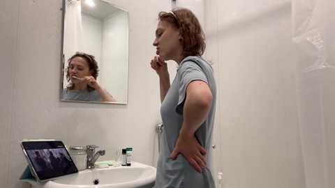 Media: Video of a woman with short brown hair, wearing a blue shirt, brushing her teeth in a small, white-tiled bathroom with a mirror, sink, and tablet on the counter.