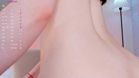 Media: A close-up video of a nude person with fair skin, showing their back and shoulder, in a dimly lit room with a standing lamp and pink wall.