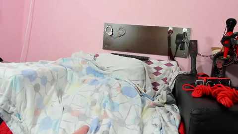 Media: Video of a pink-walled bedroom with a messy bed featuring a white duvet with pastel floral print, a black leather headboard, and a metal key hook.