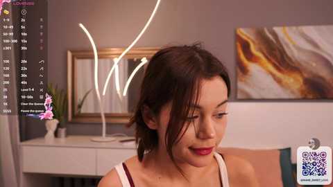 Media: Video of a young woman with dark hair and fair skin, wearing red lipstick and a white tank top, smiling slightly. Background features a modern painting, a desk with a potted plant, and a digital interface showing live stream details.