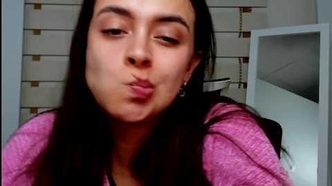 Media: Video of a young woman with long dark hair, making a kissy face, wearing a pink hoodie, in a kitchen with white cabinets and a black sink.