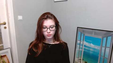 Media: A video of a young, fair-skinned woman with long, straight red hair and glasses, wearing a black dress, standing in a room with a white door and a framed painting of a beach scene.