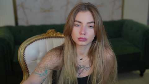 Media: Video of a young, pale-skinned woman with long, straight blonde hair, wearing a black tank top, sitting on an ornate, green velvet sofa with a white cushion. She has numerous tattoos on her shoulders and chest, and her expression is neutral.