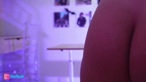 Media: Video of a close-up of a bare, light-skinned buttock with a purple glow. Background shows a white wall with framed photos and a metal table.