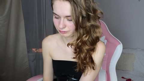 Media: Video of a young woman with fair skin, long wavy brown hair, and light makeup, wearing a strapless black dress, sitting on a pink and white gaming chair, in a dimly lit room.