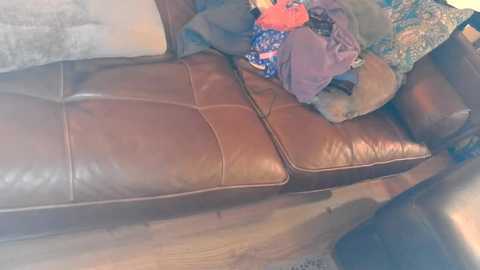 Media: Video of a brown leather couch with a plush pillow and a pile of colorful clothes, set against a beige wall and wooden floor.