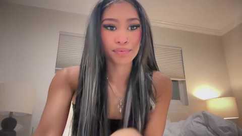 Media: Video of a young woman with long, straight black hair, wearing black clothing, in a dimly lit bedroom with beige walls, a bed, and a lamp.