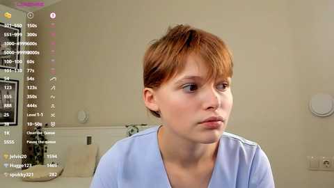 Media: Video of a red-haired, fair-skinned woman in a light blue hospital gown, looking contemplative, in a beige room with medical equipment in the background.